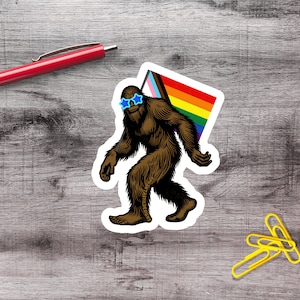 May include: A die-cut sticker featuring a brown, furry Bigfoot creature wearing bright blue star-shaped sunglasses. The creature is holding a rectangular flag with horizontal stripes in pink, white, blue, brown, black, red, orange, yellow, green, and purple.