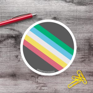 May include: A round sticker with a gray background and diagonal stripes in green, blue, white, yellow, and pink. The sticker has a white border and the text "APHANTASTICDESIGNS" at the bottom. A red pen and yellow paper clips are also visible.