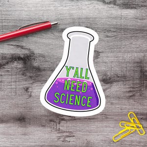 Può includere: Un adesivo bianco a forma di beuta scientifica con la scritta "Y'all Need Science" in verde. La beuta contiene liquido viola con bolle rosa. Sono visibili anche una penna rossa e delle graffette gialle.