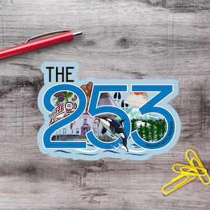 Puede incluir: Una pegatina troquelada con el texto THE 253 en una fuente azul grande. El diseño incluye ilustraciones de un Bigfoot, una orca, una montaña, un puente, una cafetería y árboles de hoja perenne. Un bolígrafo rojo y clips amarillos son visibles.