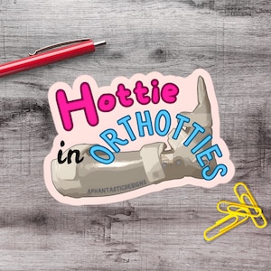May include: A die-cut sticker featuring a grey orthopaedic leg brace with the text Hottie in ORTHOTTIES in pink, black, and blue lettering. The sticker includes the small text APHANTASTICDESIGNS printed on the lower portion of the brace.