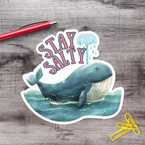 May include: A sticker featuring a blue whale swimming in blue water. The sticker has the words "STAY SALTY" in pink and white lettering. The whale is surrounded by water and a blue water spout.
