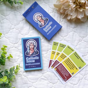 Op de afbeelding: Een blauwe en witte doos met de tekst "Catholic Trivia for Kids and Adults 1st Edition" en een afbeelding van Jezus. Er zijn ook verschillende gele en groene kaarten met vragen over het katholicisme.