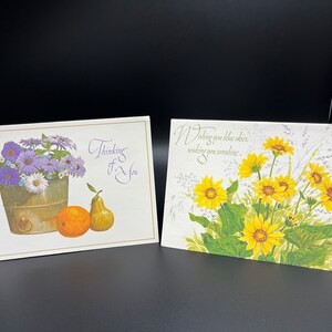 May include: Two greeting cards with floral designs. The first card features a purple and white flower arrangement in a metal bucket with an orange and a pear. The text on the card reads "Thinking of You". The second card features a bouquet of yellow flowers with green leaves and the text "Wishing you blue skies, wishing you sunshine..."
