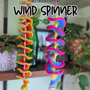 May include: Two spiral crochet wind spinners hanging vertically. The left spinner features yellow, orange, and pink yarn with a yellow tassel. The right spinner uses blue, pink, and yellow yarn with a small disco ball at the bottom. Text reads: FREE CROCHET PATTERN WIND SPINNER.