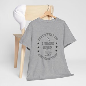Könnte beinhalten: Ein meliertes graues T-Shirt mit dem schwarzen Text "THAT'S WHAT I DO I SHAKE STUFF AND I KNOW THINGS" in einem kreisförmigen Design. Das T-Shirt hängt an einem Holzkleiderbügel. Eine weiße Baseballkappe liegt auf einem Holzstuhl.