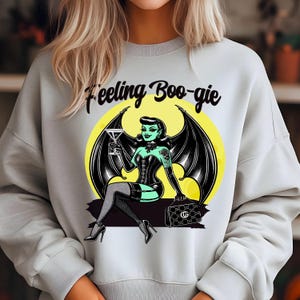 May include: A light gray sweatshirt with the text "Feeling Boo-gie" printed on the front. The graphic features a green-skinned woman with bat wings, holding a martini glass, sitting on a crescent moon.