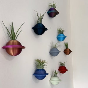 Puede incluir: Nueve macetas de diferentes colores con plantas colgadas en una pared blanca. Las macetas tienen forma de planetas y tienen un anillo alrededor. Las plantas son todas verdes y frondosas.