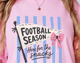 Op de afbeelding: Een lichtroze T-shirt met korte mouwen en een afbeelding van een wimpelvlag met de tekst FOOTBALL SEASON en Here for the snacks, geaccentueerd door een roze strik en verticale blauwe strepen. Het shirt wordt gedragen met een lichtblauwe spijkerbroek.