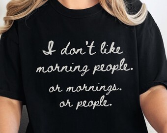 Op de afbeelding: Een zwart T-shirt met korte mouwen en witte cursieve tekst die luidt: I don't like morning people. or mornings. or people. Het shirt heeft een ronde hals en een relaxte pasvorm, gemaakt van effen zwarte stof met duidelijke letters.