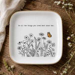 May include: A square white ceramic tray with a gold-toned rim. The center features a black line-art illustration of various wildflowers and a single orange butterfly. The text "Be all the things you loved most about her." is printed in black.