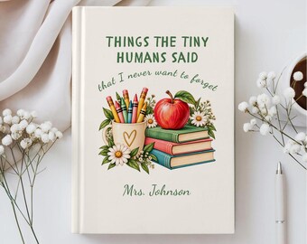 Op de afbeelding: Een wit hardcover boek met de titel Things the Tiny Humans Said that I never want to forget, voorzien van een afbeelding van een mok met kleurpotloden, een rode appel, gestapelde boeken en madeliefjes, met de naam Mrs. Johnson onderaan.