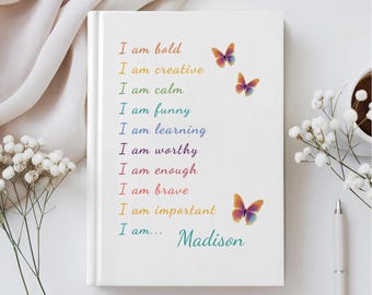 Op de afbeelding: Een wit notitieboek met harde kaft en gekleurde tekst: I am bold, I am creative, I am calm, I am funny, I am learning, I am worthy, I am enough, I am brave, I am important, I am... Madison. Er staan drie kleine vlinders op de kaft gedrukt.