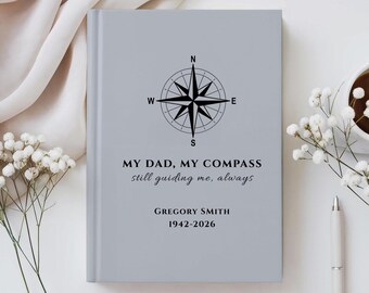 Op de afbeelding: Een lichtgrijs hardcover boek met een zwarte kompasroosafbeelding en de tekst: MY DAD, MY COMPASS, still guiding me, always, GREGORY SMITH, 1942-2026. De kaft heeft een gladde, matte afwerking met gecentreerde zwarte typografie.