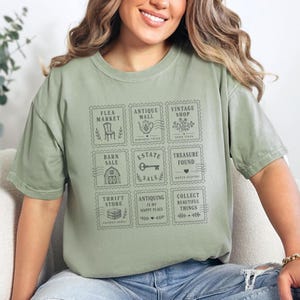 May include: A sage green short-sleeve t-shirt featuring a grid of nine rectangular stamp-like graphics. Each stamp contains black line art and text including Flea Market, Antique Mall, Vintage Shop, Barn Sale, Estate Sale, Treasure Found, Thrift Store, Antiquing Is My Happy Place, and Collect Beautiful Things.