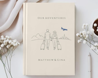 Op de afbeelding: Een crèmekleurig hardcover boek met de titel Our Adventures en de namen Matthew & Gina onder een lijntekening van een koppel dat elkaars hand vasthoudt en koffers trekt, met bergen, een stadssilhouet en een blauw vliegtuig.
