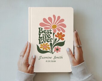 Op de afbeelding: Een crèmekleurig notitieboek met harde kaft en een bloemmotief in roze, oranje, geel en groen. De tekst Best Life Ever, Jasmine Smith en 11/01/2026 staat in een donkergroen lettertype. Het ontwerp bevat een grote roze bloem.