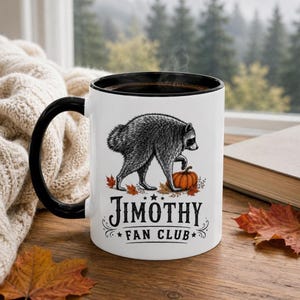 Puede incluir: Una taza de cerámica blanca con asa y borde negros, que presenta una ilustración en blanco y negro de un mapache junto a una calabaza naranja rodeada de hojas de otoño, con el texto "JIMOTHY FAN CLUB" impreso en letras negras.