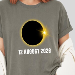 Op de afbeelding: Grijs T-shirt met een zonsverduistering ontwerp, een zwarte cirkel en een gele corona. De tekst "12 AUGUST 2026" is in het wit gedrukt. Het shirt maakt deel uit van de Lotus Sage collectie.
