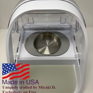 May include: A white and clear pet feeder with a stainless steel bowl. The feeder has a clear, arched top and a white base. The text "Made in USA" is visible.