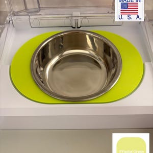 Puede incluir: Un cuenco para mascotas de acero inoxidable se encuentra sobre una alfombrilla verde pastel dentro de un comedero blanco. La alfombrilla tiene forma ovalada. Las palabras "MADE IN U.S.A." están impresas en una pegatina blanca con la bandera americana.