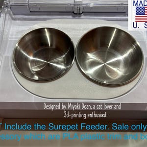 May include: Two stainless steel pet food bowls sit side-by-side on a white plastic base. The bowls are round with a shiny, reflective surface. The image includes text that says "MADE IN U.S.A."