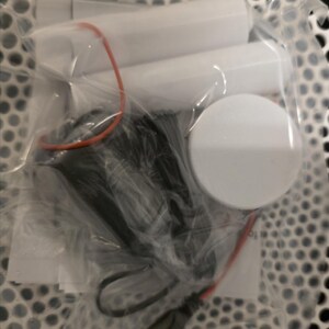 May include: A clear plastic bag containing several white cylindrical objects, a black component with a cable, and a white circular object. An orange rubber band and red wire are also visible.
