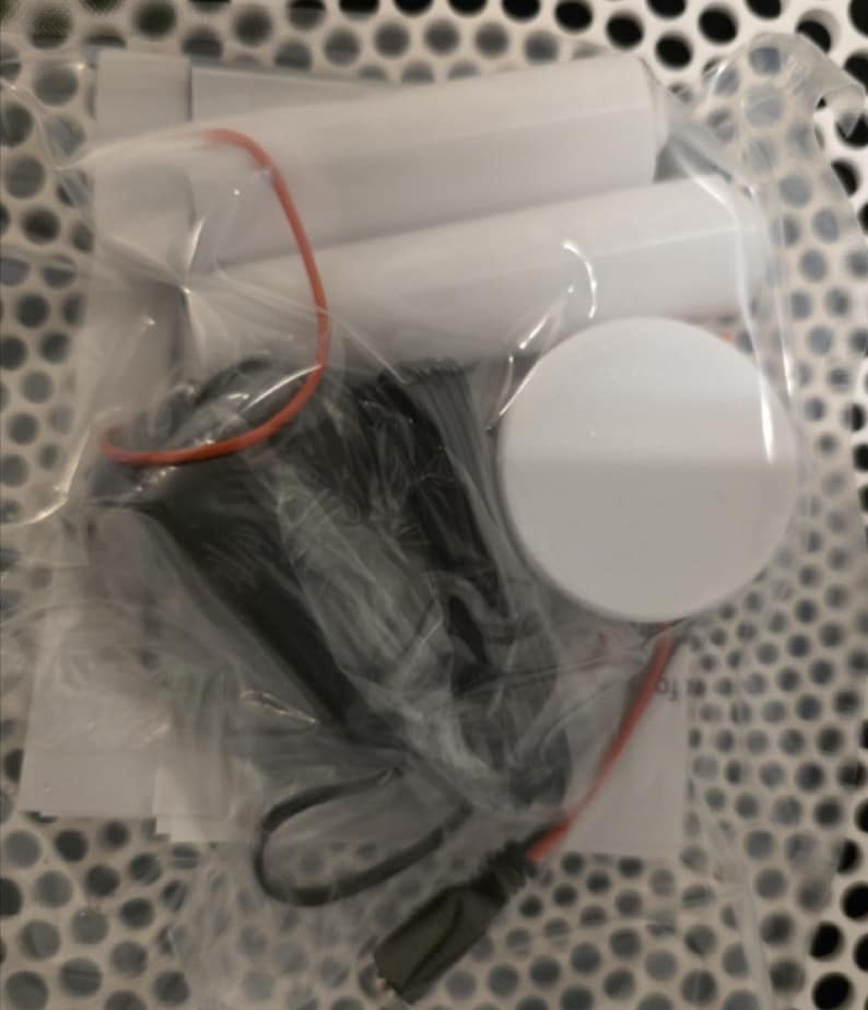 May include: A clear plastic bag containing several white cylindrical objects, a black component with a cable, and a white circular object. An orange rubber band and red wire are also visible.