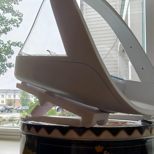 May include: A white and clear plastic object with a unique design, possibly a kitchen appliance or a decorative item. It is supported by a white stand and sits atop a black and gold tin. The object has a curved, aerodynamic shape.