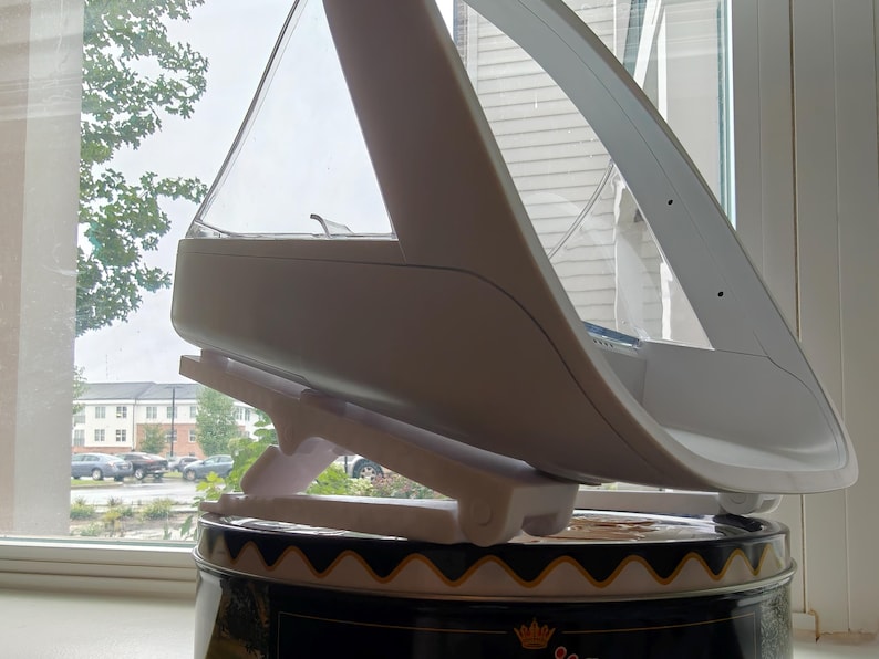 May include: A white and clear plastic object with a unique design, possibly a kitchen appliance or a decorative item. It is supported by a white stand and sits atop a black and gold tin. The object has a curved, aerodynamic shape.