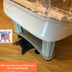 Puede incluir: Un comedero blanco y transparente para mascotas con una base gris impresa en 3D. Un adorno negro con forma de gato está debajo del comedero. La imagen incluye una pegatina "Made in U.S.A." y el texto "Sale only 3D printed titled base, not included feeder."