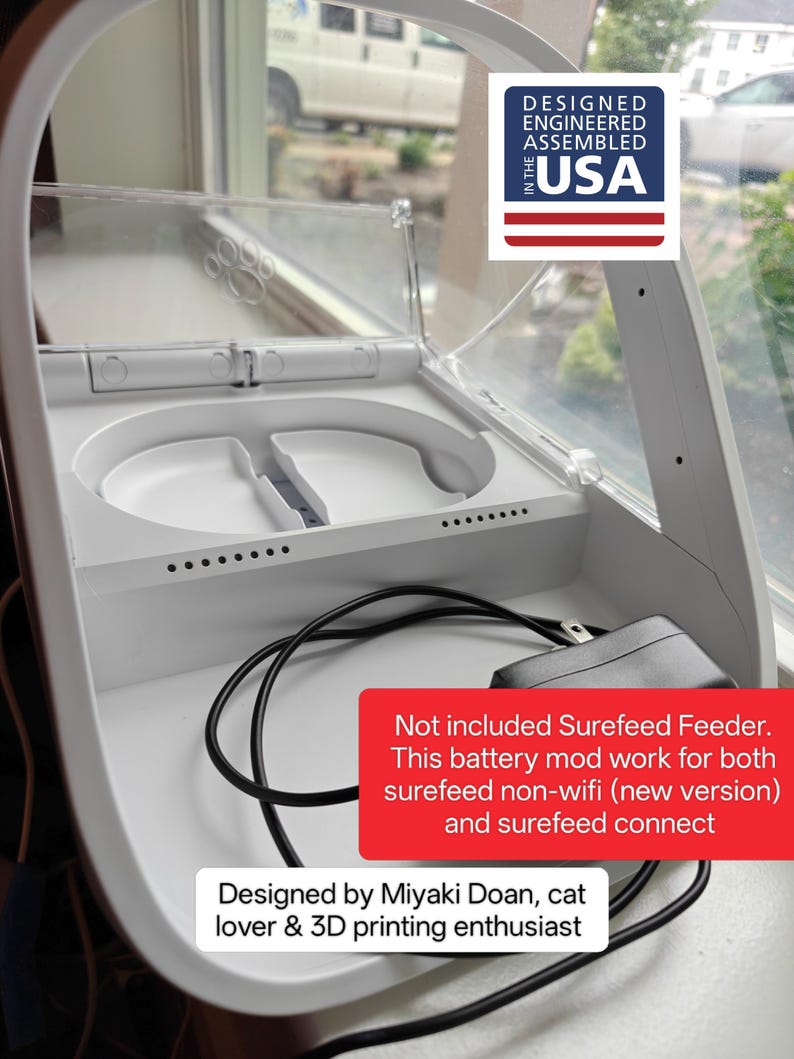 May include: White pet food feeder with a clear lid, designed and assembled in the USA. The feeder has two food compartments and a power cord. The text on the image says it is a battery mod for Surefeed feeders.