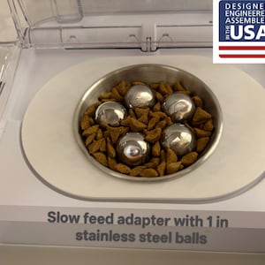 Puede incluir: Un dispensador de comida para mascotas blanco con un cuenco de acero inoxidable que contiene comida seca y cuatro bolas de acero inoxidable. El dispensador tiene una tapa transparente y el texto "Slow feed adapter with 1 in stainless steel balls".