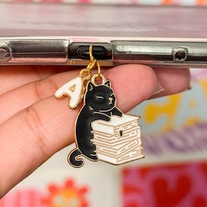 May include: A black cat charm with a gold letter 'A' and a stack of papers. The charm is attached to a phone case.