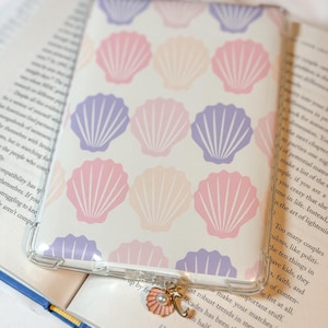 May include: A clear phone case featuring a repeating pattern of pastel pink, peach, and purple seashells. A small charm with a pink seashell and pearl hangs from the case. The case rests on an open book.