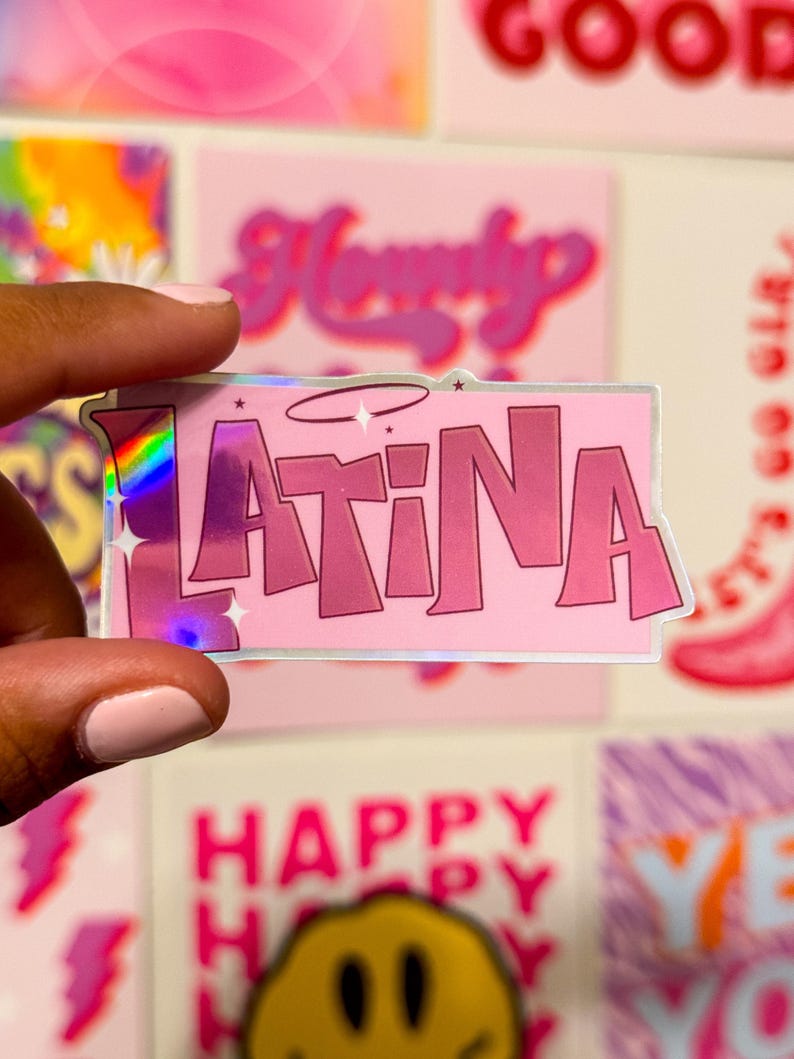 Puede incluir: Una pegatina hologr&aacute;fica rosa y plateada con la palabra "LATINA" en letras rosas con un halo sobre la palabra. La pegatina est&aacute; siendo sostenida por una mano.