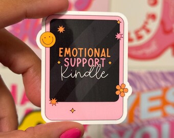 May include: A pink sticker with the words "EMOTIONAL SUPPORT Kindle" in orange and white. The sticker has a black background and is decorated with a yellow smiley face, orange stars, and a flower.
