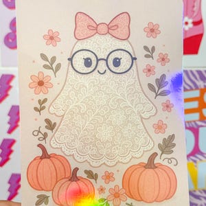May include: A rectangular sticker featuring a central ghost character with a lace-like floral pattern, wearing round black glasses and a pink bow. The ghost is surrounded by small pink flowers, green leaves, and three orange pumpkins at the base.