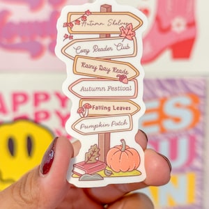 May include: A sticker featuring a signpost design with autumn-themed text. The signpost has labels that read "Autumn Shelves," "Cozy Reader Club," "Rainy Day Reads," "Autumn Festival," "Falling Leaves," and "Pumpkin Patch." A small pumpkin and book are at the bottom.