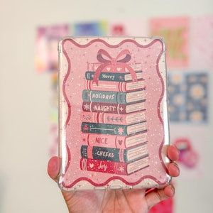 May include: A clear phone case with a pink border and a stack of books design. The books are labeled "Merry", "Holidays", "Naughty", "Nice", "Cheers", and "Joy". A pink bow is at the top of the stack.