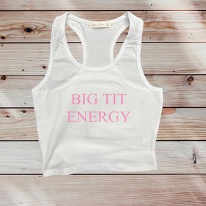 May include: White racerback crop top with the text "BIG TIT ENERGY" in pink. The top has a scoop neck and a racerback design. The shirt is made of a soft, lightweight material.