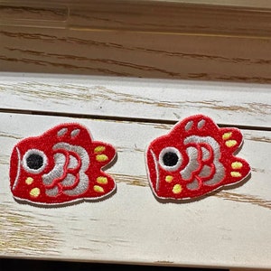 May include: Two red and white embroidered fish patches with yellow accents. The fish have black eyes and are in a cartoon style.