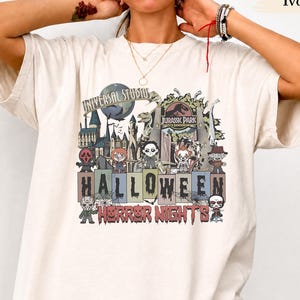 May include: An ivory short-sleeve t-shirt featuring a graphic print of Universal Studios Halloween Horror Nights. The design includes various horror characters, a Jurassic Park gate, and the text "Universal Studios" and "Halloween Horror Nights."