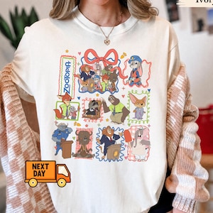 May include: An ivory crewneck t-shirt featuring a colourful graphic print of Zootopia characters arranged in various frames. The design includes Nick Wilde, Judy Hopps, Flash, and other movie figures, with a red bow at the top and small hearts.