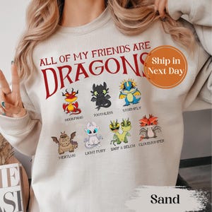 May include: Sand-colored sweatshirt featuring the text "ALL OF MY FRIENDS ARE DRAGONS" in red, with various cartoon dragon illustrations below. The sweatshirt includes the names of the dragons: Hookfang, Toothless, Stormfly, Meatlug, Light Fury, Barf & Belch, and Cloudjumper.