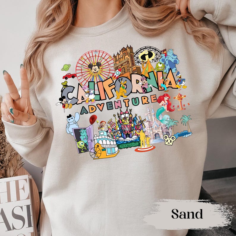 May include: Sand-colored sweatshirt featuring a colorful graphic of Disneyland characters and attractions. The design includes the text "CALIFORNIA ADVENTURE" with images of Mickey Mouse, a Ferris wheel, and other iconic park elements.