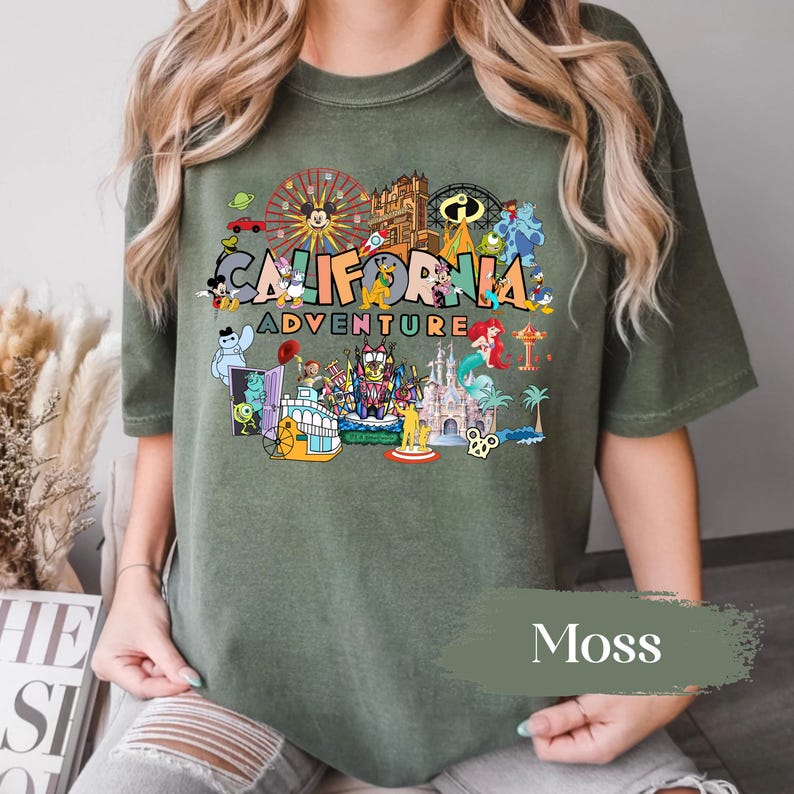 May include: Moss green t-shirt featuring a colorful graphic of California Adventure theme park attractions and characters. The text "CALIFORNIA ADVENTURE" is displayed prominently. The shirt is a relaxed fit.