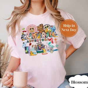 May include: Light pink t-shirt featuring a colorful graphic of the California Adventure theme park. The graphic includes various cartoon characters, a Ferris wheel, and the words "California Adventure".