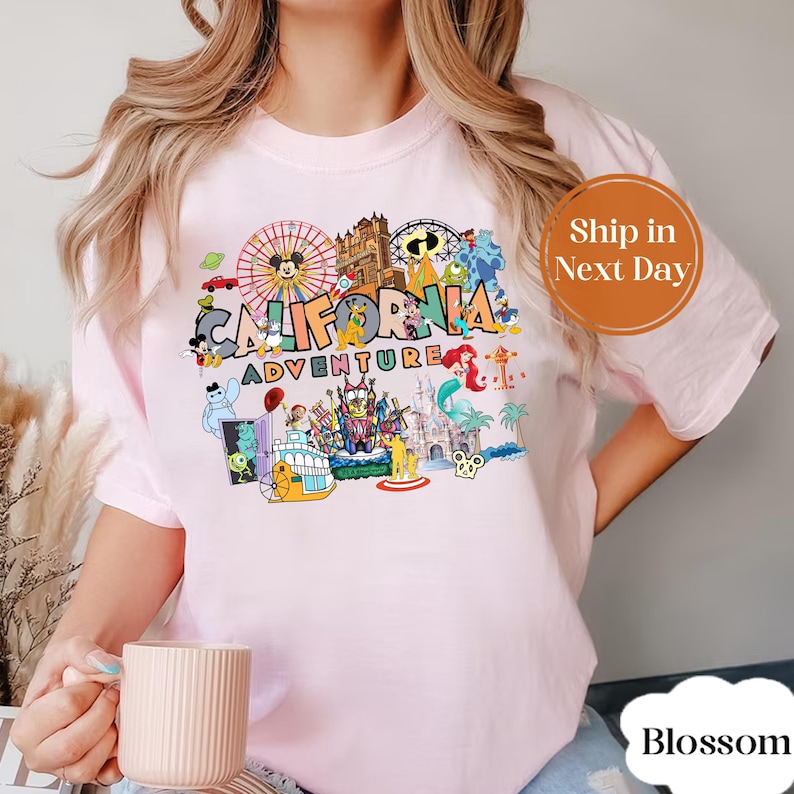 May include: Light pink t-shirt featuring a colorful graphic of the California Adventure theme park. The graphic includes various cartoon characters, a Ferris wheel, and the words "California Adventure".