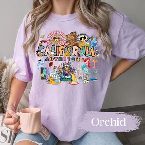 May include: A light purple t-shirt with a colorful graphic featuring the words "California Adventure" and various cartoon characters and landmarks. The shirt has short sleeves and a relaxed fit. The word "Orchid" is printed on the bottom right.